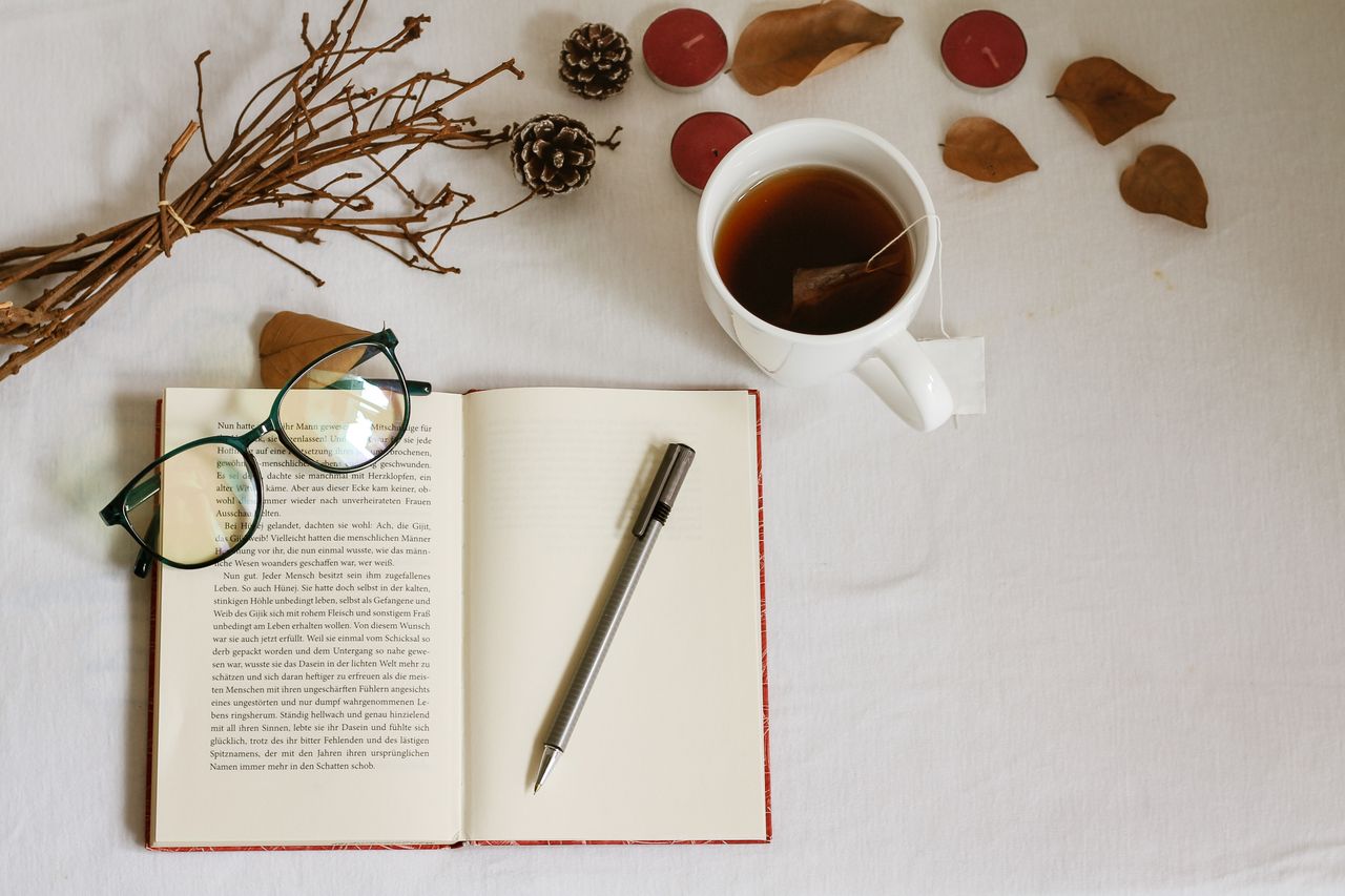 Flat lay of open book, eyeglasses, cup of tea, and autumn leaves, creating a warm, cozy reading atmosphere.